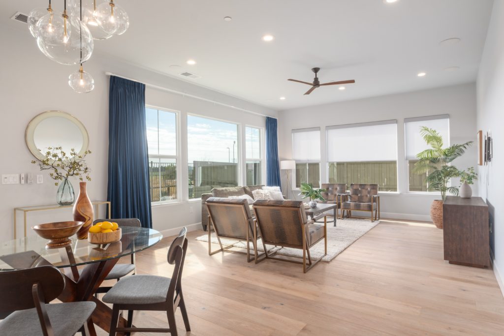 Dining chair in foreground and living room in the background with chairs and a sofa. There are large windowns flanked by blue curtains on the left side.