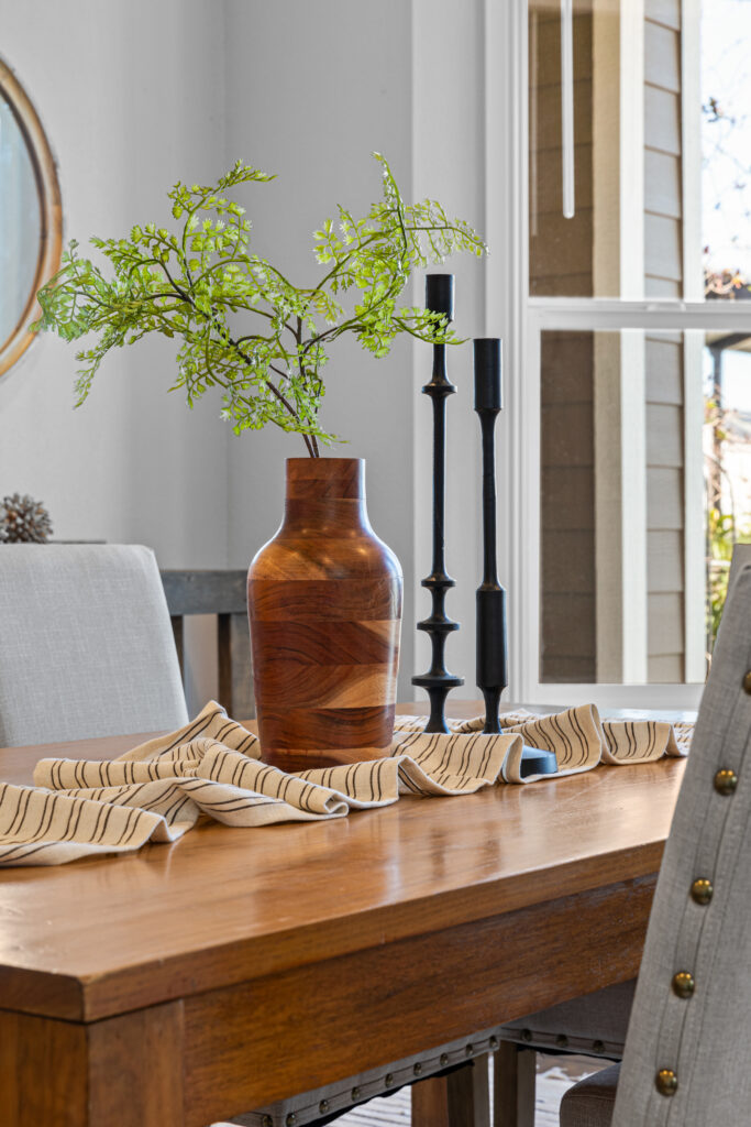 Tablescape of a wooden vase with greenery, two tall modern candlesticks sitting on a wooden table with a scrunched-up fabric runner for texture.