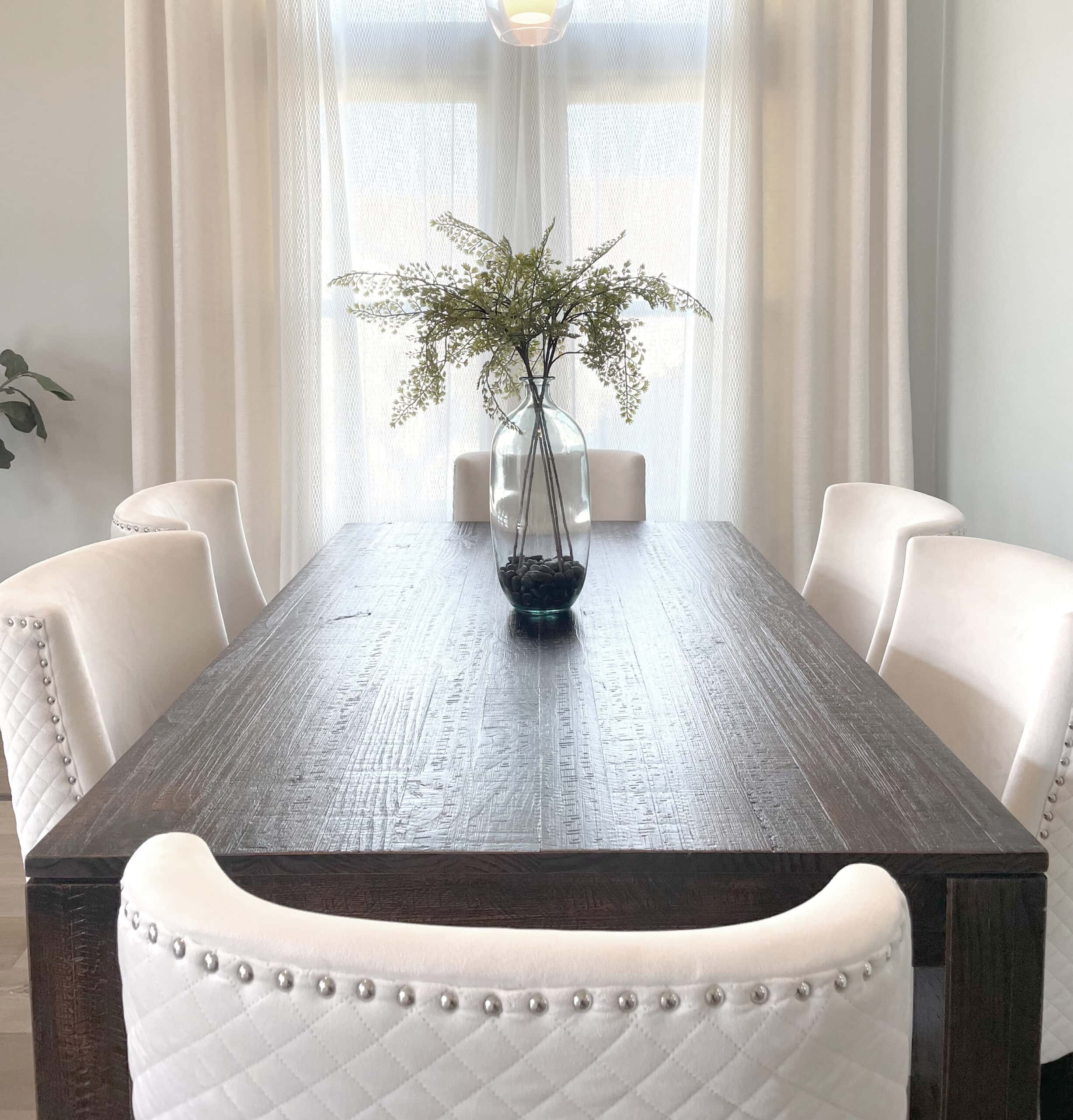 Dining table with six white chairs with diamond-back tufting. Table haas a centerpiece of a glass vase wtih greenery extending. In the background there is a window with open drapery.